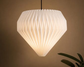 May include: A white pendant light featuring a geometric, pleated paper shade with a diamond-like silhouette. The light fitting is suspended from a thin black cord, emitting a soft, warm glow through the folded material structure.