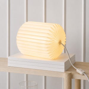 May include: A cylindrical table lamp with a pleated, off-white shade that emits a warm, soft glow. The lamp features a horizontal ribbed texture and is connected to a white power cord with an inline switch, resting on a stack of white books.