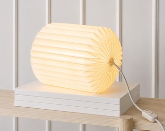 May include: A cylindrical table lamp with a pleated, off-white paper shade that emits a warm glow. The lamp features a white power cord with an inline switch and rests horizontally on a stack of two white rectangular books.