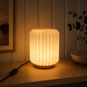 May include: A cylindrical table lamp featuring a white, accordion-folded paper shade that emits a warm, diffused glow. The lamp is supported by a circular light-toned wooden base and includes a black power cord with an inline rocker switch.