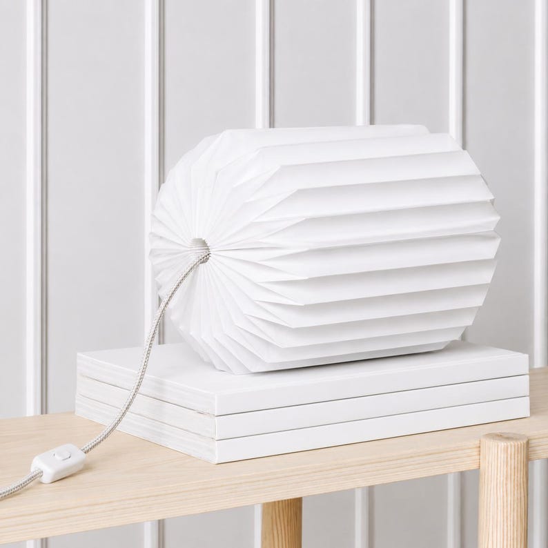 May include: A white cylindrical lamp with a pleated paper shade resting on a stack of three white books. A thin, light-colored fabric-covered power cord with a small white plastic switch extends from the center of the lamp shade.