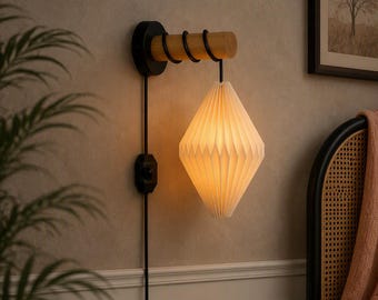 May include: A wall-mounted lamp featuring a cylindrical light-coloured wood arm with black metal accents and a black circular base. A diamond-shaped, pleated white paper lampshade hangs from the arm, emitting a warm, soft glow.