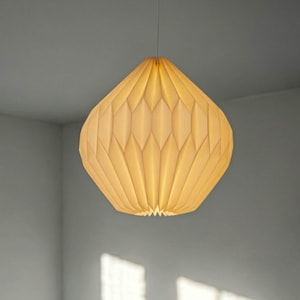 Op de afbeelding: Een crèmekleurige hanglamp met een geplooide, geometrische kap van papierachtig materiaal. De lamp heeft een ronde, druppelvormige vorm met verticale plooien die een ruitpatroon creëren. De lamp verspreidt een warme, zachte gloed van binnenuit.