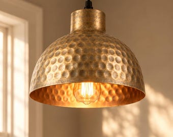 May include: A dome-shaped pendant light fitting with a hammered, antique gold-toned metal finish. The lamp features a central Edison-style light bulb that emits a warm glow, suspended from a black cord attached to the top of the shade.