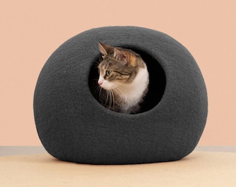 May include: A dark grey, rounded, felted wool cat cave with a circular opening. A brown and white tabby cat with green eyes is positioned inside the structure, looking towards the left side of the frame, with its head visible in the opening.