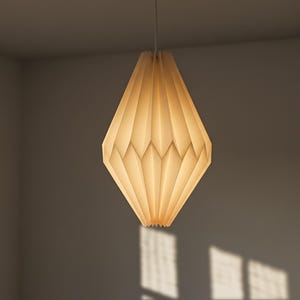 May include: A diamond-shaped pendant light fixture with a pleated, off-white paper shade. The structure features a geometric, folded design that emits a soft, warm glow from within, suspended by a thin white cord from a ceiling mount.