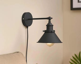 May include: A matte black wall-mounted lamp featuring a circular base, a horizontal arm, and a conical shade. A black power cord hangs from the base, and a single light bulb is visible within the shade, casting a soft glow downwards.