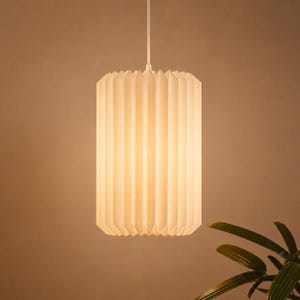 Puede incluir: Una lámpara colgante cilíndrica con una pantalla blanca de pliegues verticales que emite un brillo suave y cálido. La lámpara está suspendida de un cable blanco fino sujeto en el centro superior, creando una difusión de luz uniforme.
