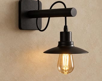 May include: A matte black wall-mounted lamp featuring a square base plate, a horizontal cylindrical arm, and a curved cord. A wide, shallow conical shade hangs from the arm, housing a clear glass Edison-style light bulb with visible filaments.