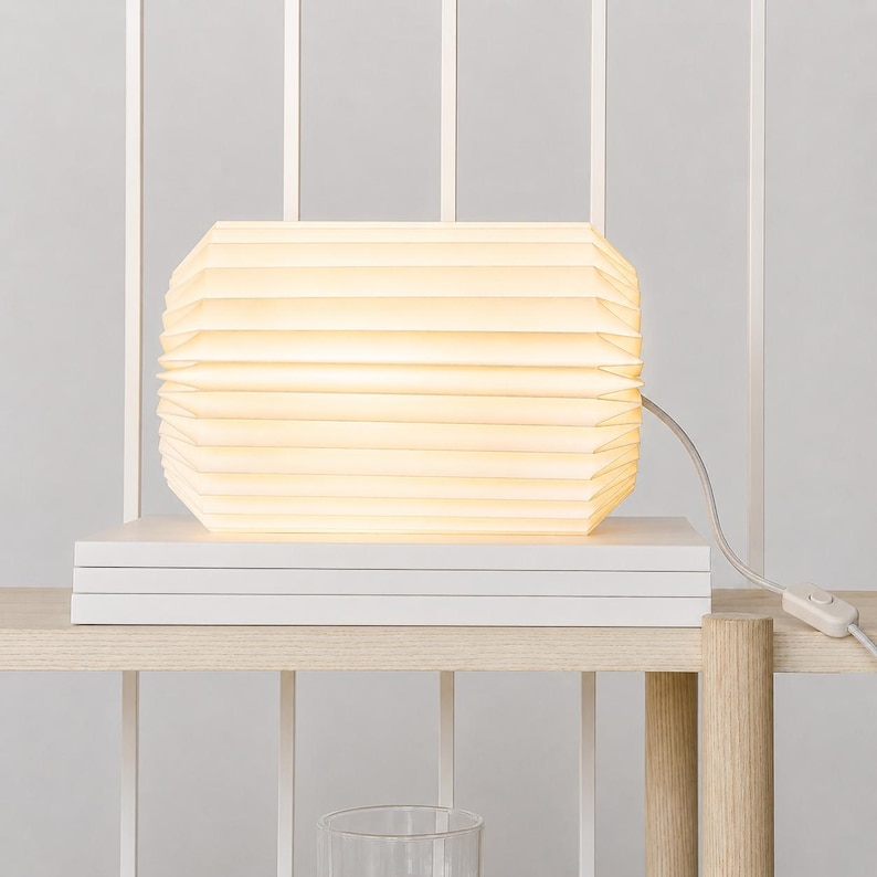 May include: A horizontal, cylindrical table lamp with a pleated, off-white paper shade that emits a warm, diffused glow. The lamp rests on a stack of white rectangular books and includes a white power cord with an inline toggle switch.