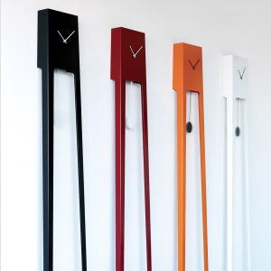 Op de afbeelding: Vier moderne, minimalistische wandklokken in zwart, rood, oranje en wit. Elke klok heeft een rechthoekige bovenkant met een wijzerplaat en lange, slanke poten. De oranje klok heeft een zwarte slinger. De klokken zijn aan een witte muur bevestigd.