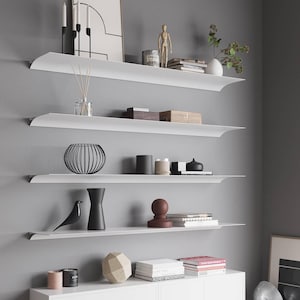 Può includere: Una moderna mensola a parete con quattro ripiani bianchi. I ripiani espongono vari oggetti decorativi, tra cui candele, vasi, libri e sculture. Lo sfondo è una parete grigia uniforme, che crea un'estetica minimalista.