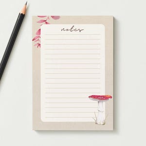 May include: A rectangular notepad with a beige background, horizontal lines, and the word notes printed at the top. The design features a watercolour illustration of a red and white mushroom in the corner and pink leaves at the top left.
