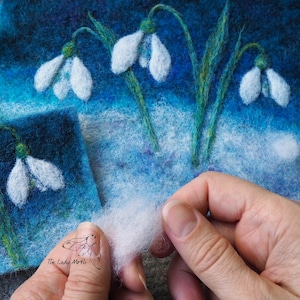 Op de afbeelding: Een close-up van een hand die een stuk witte wolvezels vasthoudt. De vezel wordt weggetrokken van een blauw en wit viltkunstwerk dat sneeuwklokjes voorstelt. Het kunstwerk is gesigneerd met "The Lady Moth".