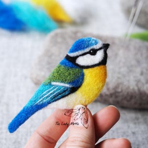 Może przedstawiać: Niebieski ptak z żółtym brzuchem, zielonymi plecami i niebieskimi skrzydłami, wykonany z filcu. Ptak siedzi na palcu.