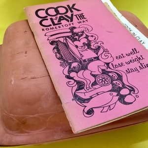 May include: A pink cookbook titled "Cook in Clay the Romertopf Way" with illustrations of food and the text "eat well lose weight stay slim".