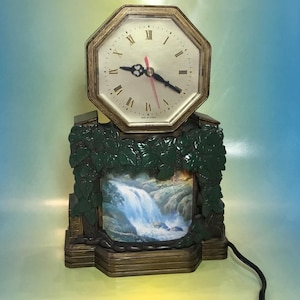 May include: A vintage clock with a waterfall scene. The clock has a green and brown base with a picture of a waterfall in the center. The clock face is octagonal and has Roman numerals. The clock is plugged in and has a cord.