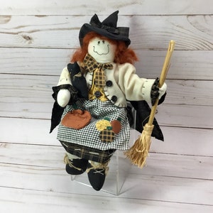 May include: A fabric Halloween witch doll with red hair, a black hat, and a cream-colored jacket. The doll has a black cat, a pumpkin-shaped bag, and a broom. The witch is wearing a black and white checkered skirt and black shoes.