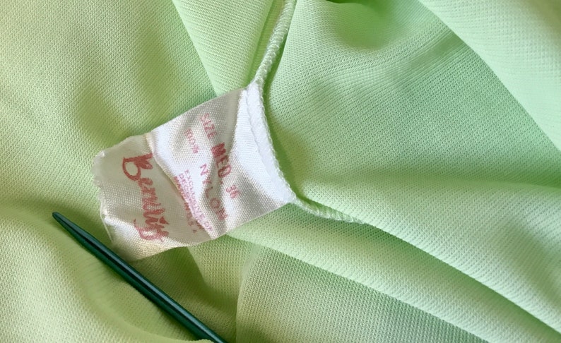 May include: A white tag with the text "Beauty" in red lettering and "Size Med 36 100% Nylon" in black lettering. The tag is attached to a light green fabric.