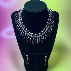 May include: A vintage necklace and earring set displayed on a black bust. The necklace features a gold chain with alternating small white beads and dangling dark red teardrop-shaped beads. The matching earrings have a similar design, with a single teardrop bead.