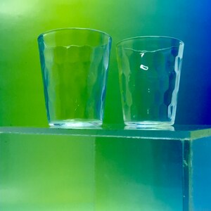 May include: Two clear glass tumblers with a textured surface. The glasses are sitting on a green and blue surface.