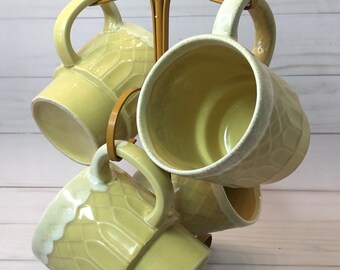 May include: Set of six yellow ceramic mugs with a textured design, displayed on a gold metal mug rack.