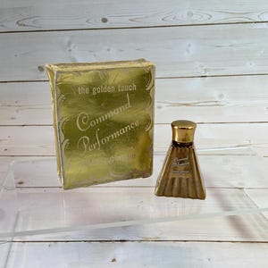 May include: A vintage perfume bottle and box set. The box is gold with the words "the golden touch Command Performance" in elegant script. The perfume bottle is a small, gold-toned triangular shape with a gold cap. The set is displayed on a clear acrylic surface.