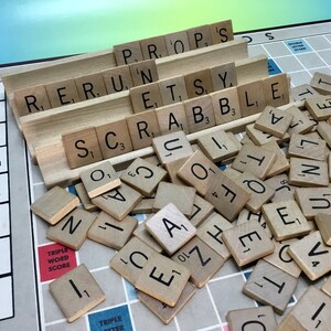 May include: A close-up of a Scrabble game in progress. The tiles are scattered on the board, with some tiles still in the rack. The tiles spell out the word "SCRABBLE" and other words like "PROP'S", "RERUN", and "ETSY".
