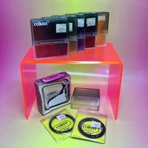 May include: A collection of Cokin camera filters and accessories. The image features several filters in clear plastic cases, a box, and two circular filters in yellow packaging. The filters are arranged on a neon orange acrylic shelf.