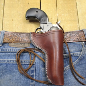May include: A silver and black handgun in a brown leather holster, attached to a blue denim jeans with a brown leather belt. The holster is stitched with white thread and has a leather tie. The belt has a decorative pattern.