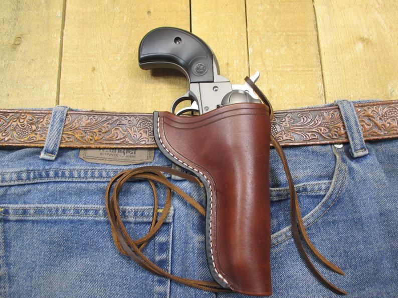 May include: A silver and black handgun in a brown leather holster, attached to a blue denim jeans with a brown leather belt. The holster is stitched with white thread and has a leather tie. The belt has a decorative pattern.