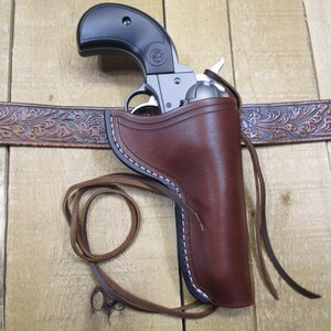 May include: A brown leather holster with white stitching holds a silver and black revolver. The holster is attached to a brown, embossed leather belt. A leather strap is tied to the holster.