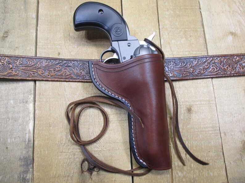 May include: A brown leather holster with white stitching holds a silver and black revolver. The holster is attached to a brown, embossed leather belt. A leather strap is tied to the holster.