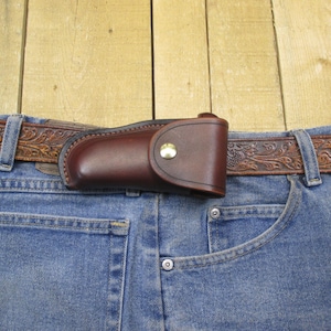 Può includere: Una cintura in pelle marrone con un motivo floreale in rilievo e un fodero per coltello in pelle marrone attaccato. La cintura è indossata sopra un paio di jeans blu.