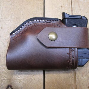 May include: A brown leather holster with a black handgun inside. The holster has a decorative stitched design along the top edge and a strap with a brass button closure. The holster is designed to hold a handgun.