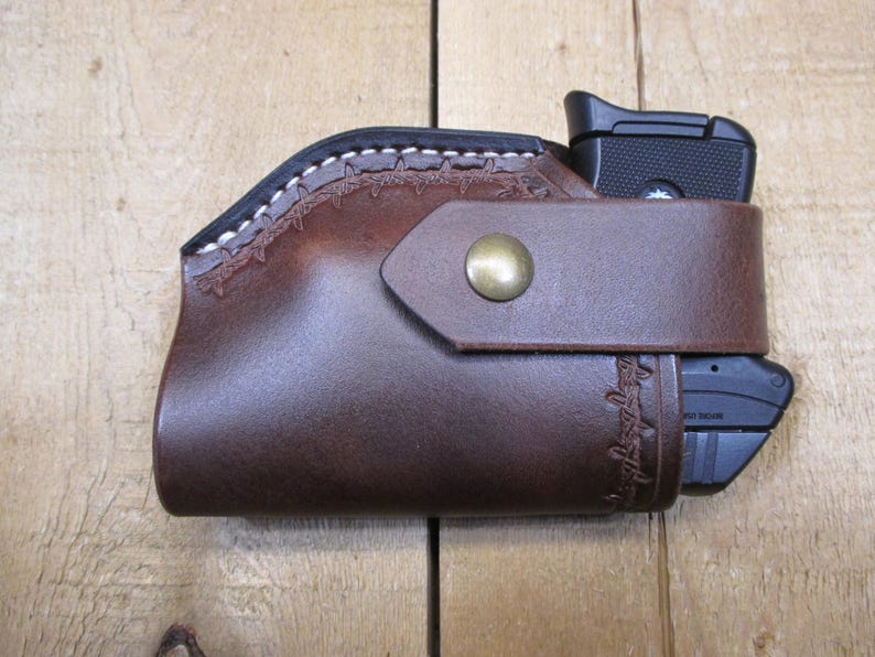 May include: A brown leather holster with a black handgun inside. The holster has a decorative stitched design along the top edge and a strap with a brass button closure. The holster is designed to hold a handgun.