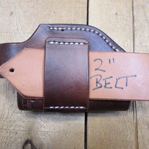 May include: A brown leather holster with white stitching, designed to fit a 2-inch belt. The holster is made of sturdy leather and features a loop for belt attachment. The text "2" and "BELT" is handwritten on a tan leather piece.