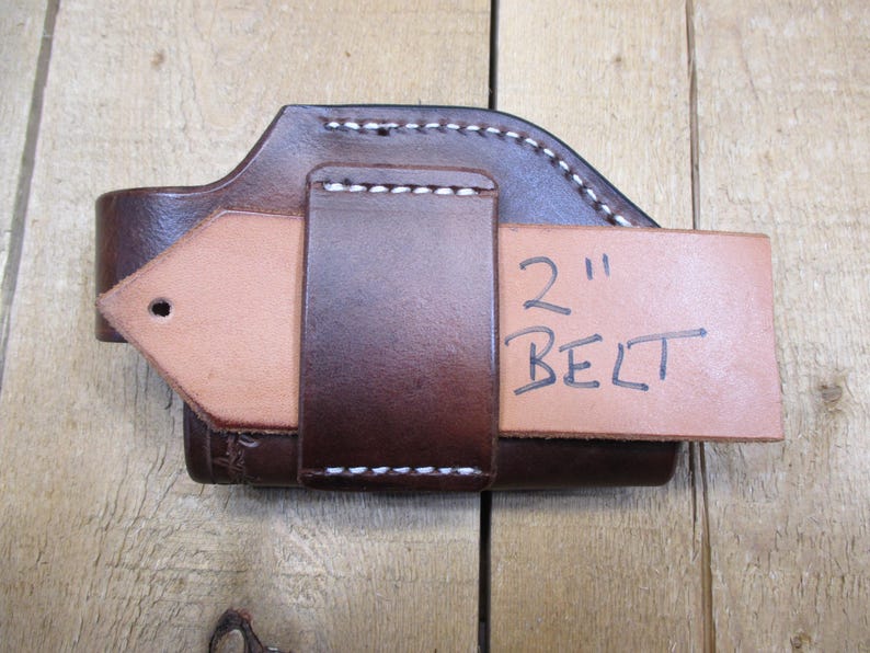 May include: A brown leather holster with white stitching, designed to fit a 2-inch belt. The holster is made of sturdy leather and features a loop for belt attachment. The text "2" and "BELT" is handwritten on a tan leather piece.