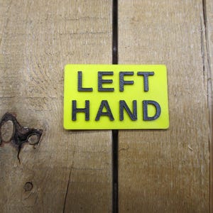 May include: A rectangular, neon yellow sign with the words "LEFT HAND" in black, block letters. The sign is set against a wooden background with visible grain and knots.