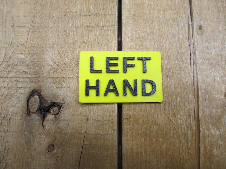 May include: A rectangular, neon yellow sign with the words "LEFT HAND" in black, block letters. The sign is set against a wooden background with visible grain and knots.