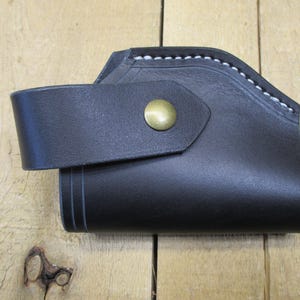 May include: Black leather holster with a curved design and a strap secured by a brass button. White stitching accents the top edge. The holster is likely for a small firearm or tool.