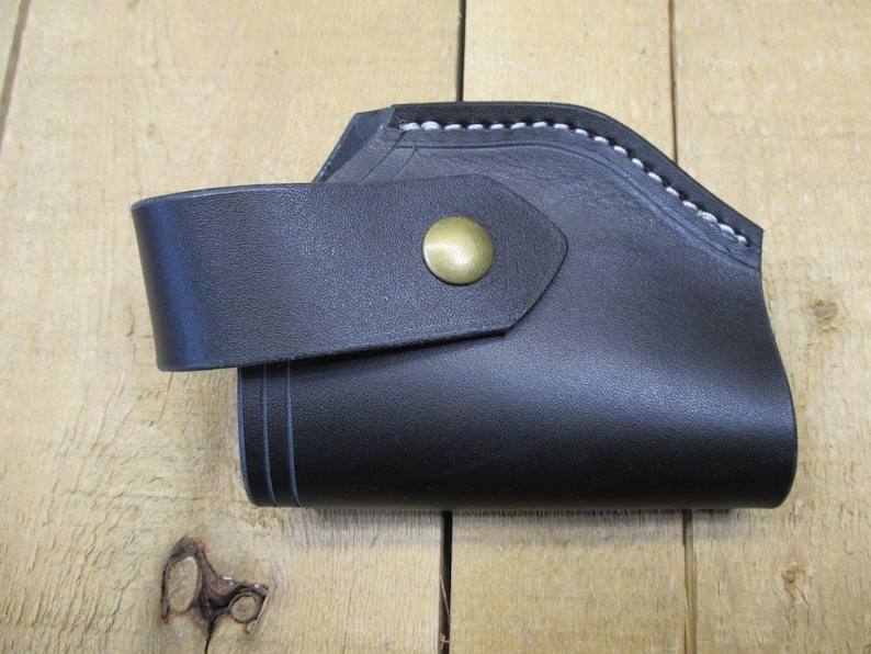May include: Black leather holster with a curved design and a strap secured by a brass button. White stitching accents the top edge. The holster is likely for a small firearm or tool.