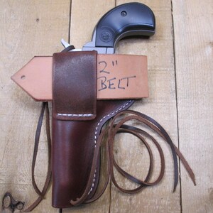 May include: A brown leather holster with white stitching, designed to hold a small handgun. The holster is attached to a light brown leather belt loop. The text "2" BELT" is handwritten on the belt loop.