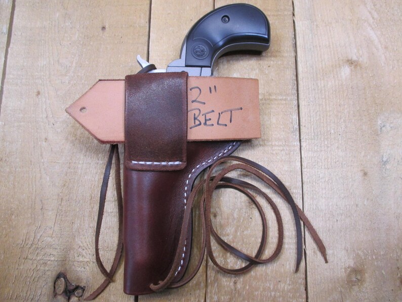 May include: A brown leather holster with white stitching, designed to hold a small handgun. The holster is attached to a light brown leather belt loop. The text "2" BELT" is handwritten on the belt loop.