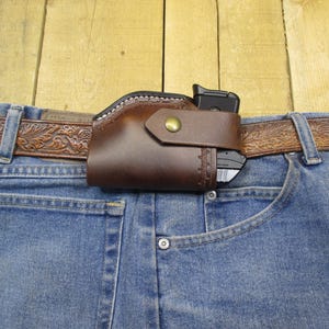 May include: A brown leather holster with a snap closure, holding a black handgun, attached to a brown, embossed leather belt. The belt is worn with blue denim jeans.