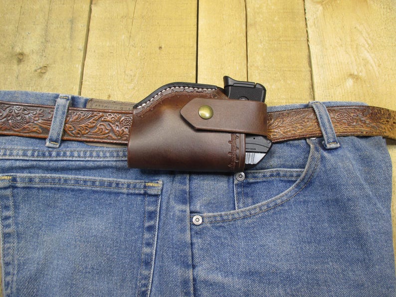 May include: A brown leather holster with a snap closure, holding a black handgun, attached to a brown, embossed leather belt. The belt is worn with blue denim jeans.