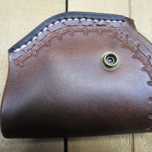 May include: A brown leather holster with a decorative barbed wire design. The holster has a black top edge with white stitching and a brass-colored snap. The leather is a rich, dark brown color.