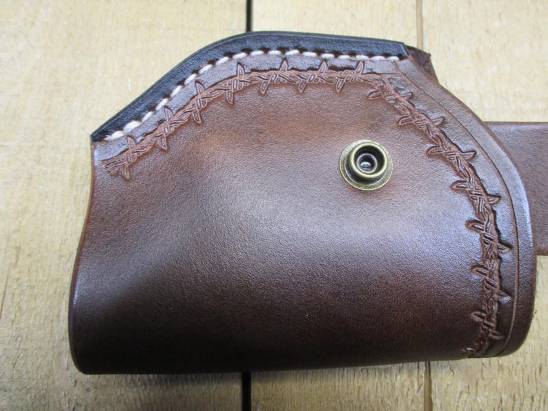 May include: A brown leather holster with a decorative barbed wire design. The holster has a black top edge with white stitching and a brass-colored snap. The leather is a rich, dark brown color.