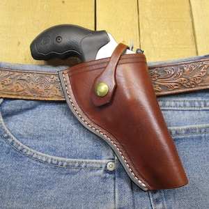 Può includere: Un fodero in pelle marrone con chiusura a scatto, progettato per contenere un'arma da fuoco. Il fodero è indossato su una cintura sopra un paio di jeans blu.