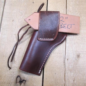 May include: A brown leather holster with white stitching and a belt loop. The holster is designed to fit a 2-inch belt. The image shows the holster on a wooden surface.